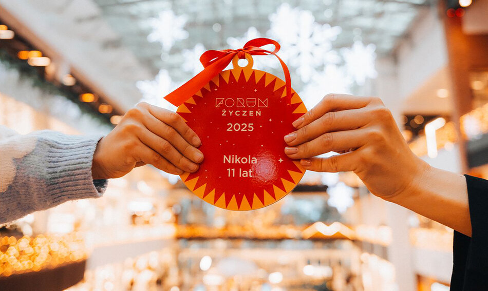 Dwie ręce trzymają papierową bombkę z napisanym imieniem dziecka i jego wiekiem oraz napisem Forum Życzeń.