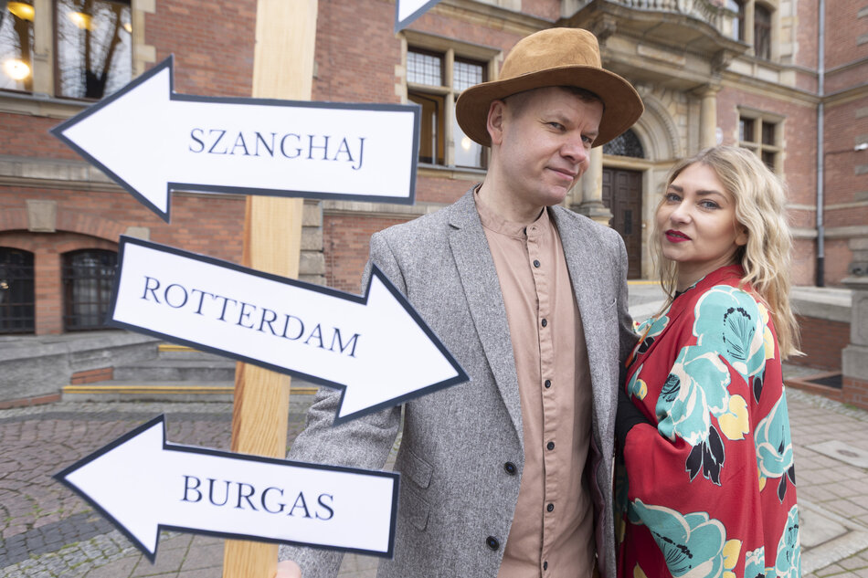 Na zdjęciu widać dwie osoby stojące obok drogowskazu z nazwami miast: Szanghaj, Rotterdam i Burgas, ustawionego na tle ceglanego budynku. Stylizowane stroje oraz układ kadrów sugerują motyw podróży i symbolicznego wyboru kierunku. 
