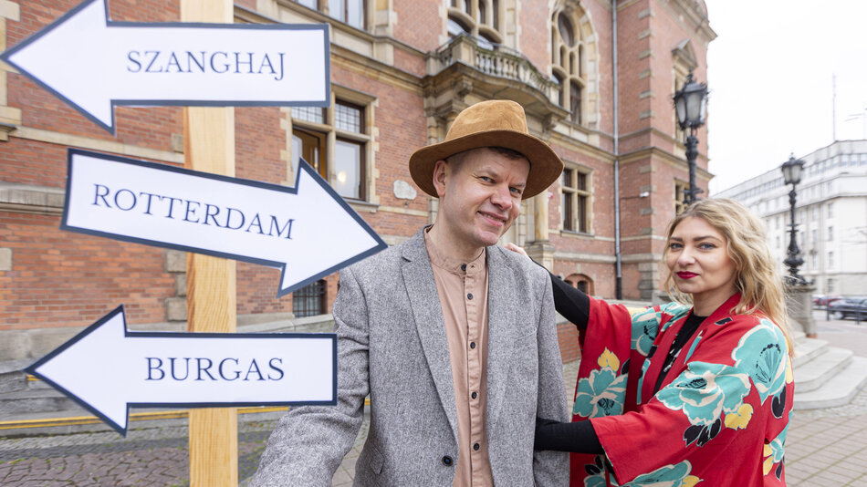Na zdjęciu widać dwie osoby stojące obok drogowskazu z nazwami miast: Szanghaj, Rotterdam i Burgas, ustawionego na tle ceglanego budynku. Stylizowane stroje oraz układ kadrów sugerują motyw podróży i symbolicznego wyboru kierunku. 