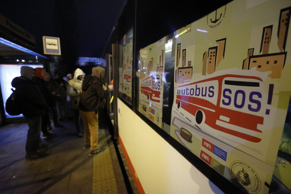 Autobus miejski stoi na przystanku wieczorem, do drzwi stoi kolejka ludzi, na oknie jest plakat z napisem Autobus SOS.