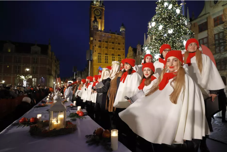 Świąteczne spotkanie na Długim Targu w Gdańsku. Grupa osób w białych pelerynach i czerwonych czapkach stoi przy długim, udekorowanym stole na tle oświetlonego Ratusza i choinki.