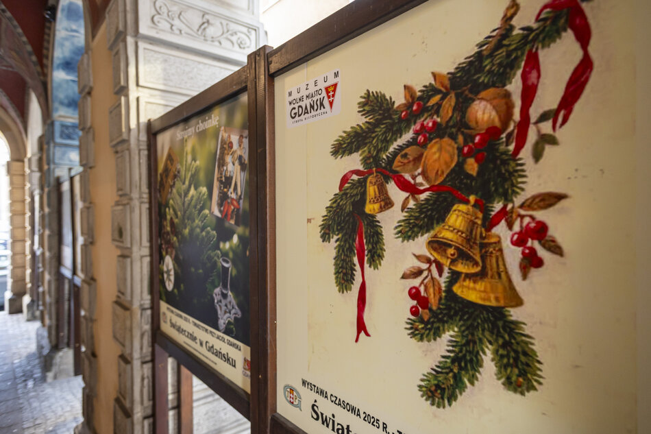 Na zdjęciu widoczna jest ekspozycja plenerowa w arkadach muzeum, z planszami informacyjnymi poświęconymi tematyce świątecznej. Na pierwszym planie znajduje się ilustracja gałązki świerkowej z dzwonkami i wstążkami, umieszczona w ramie wystawowej. 