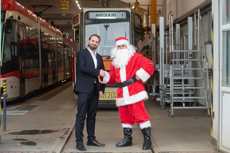 Mężczyzna w garniturze ściska dłoń człowiekowi przebranemu za mikołaja. Stoją w zajezdni tramwajowej przed tramwajem. 