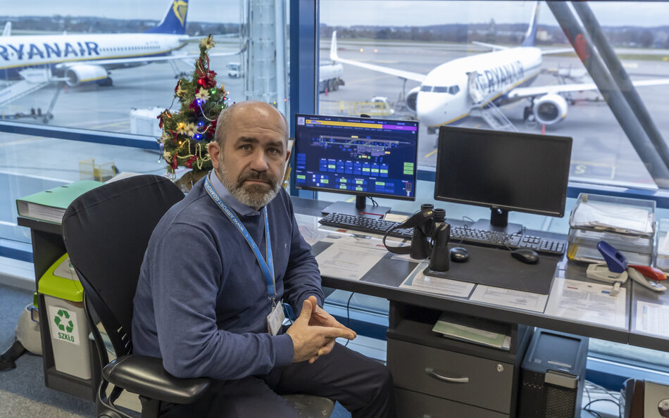 Mężczyzna siedzi na fotelu przy pulpicie z ekranami, za nim przez okno widać lotnisko i samoloty.