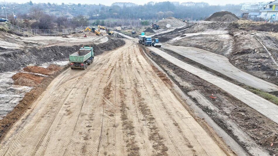 na zdjęciu plac budowy nowej drogi, widać gruntową nawierzchnię i jadące po niej ciężarówki