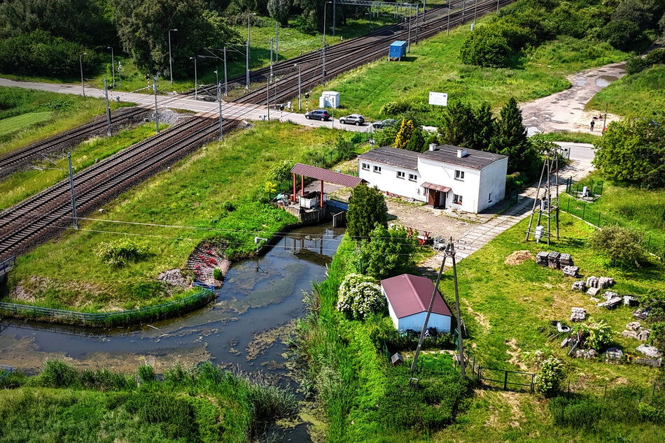 Zdjęcie z lotu ptaka na nieduży budynek przepompowni, przy której płynie rzeka, widać też tory kolejowe.