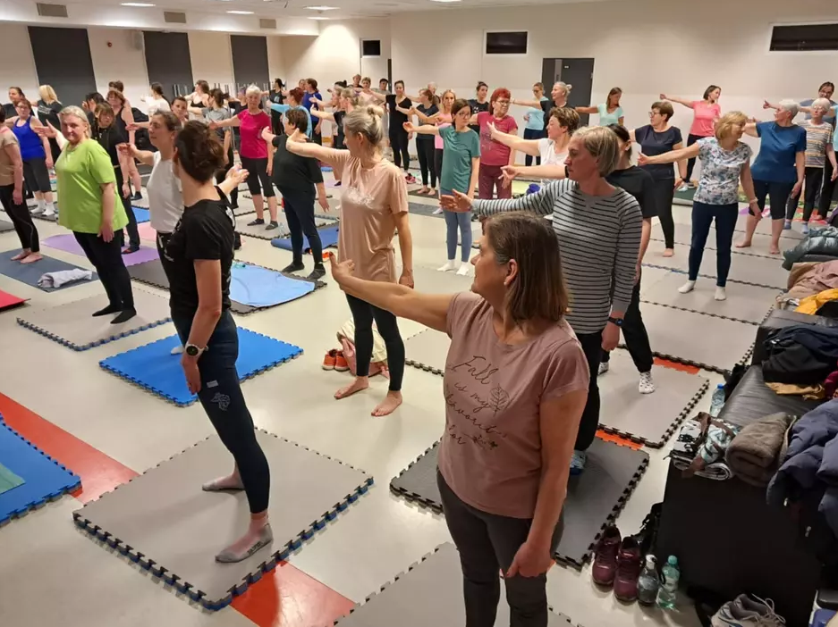 na zdjęciu kilkanaście ćwiczących osób w sali gimnastycznej, stoją i układają odpowiednio ręce