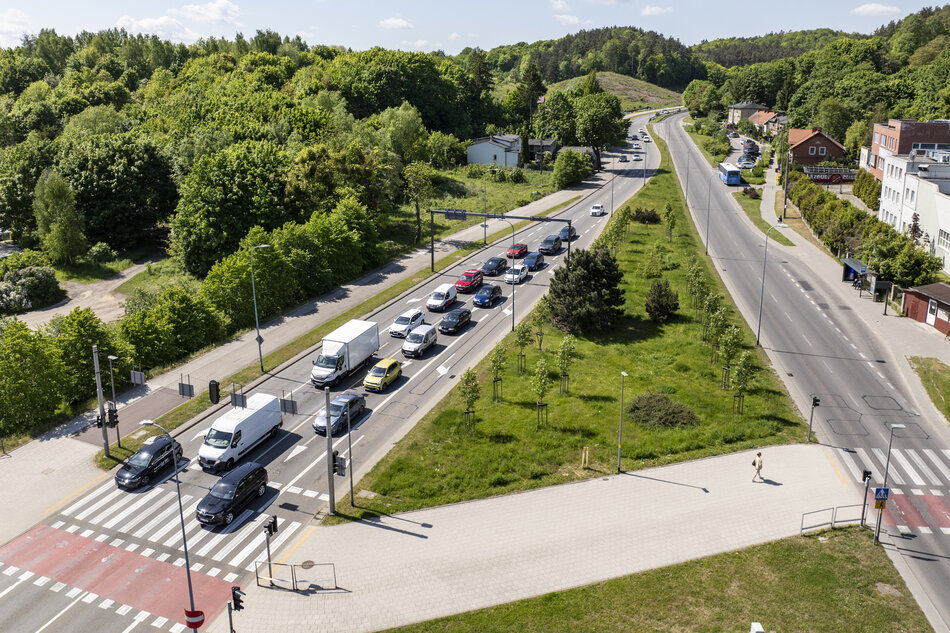 widok z lotu ptaka na ruchliwą ulicę z wieloma pasami ruchu. Ulica jest wyłożona drzewami i domami po obu stronach, a po prawej stronie znajduje się kilka budynków jest przejściem dla pieszych.Osobę można zobaczyć spacerującą po chodniku w pobliżu skrzyżowania. W tle niebo jest niebieskie, i drzewa są zielone, co wskazuje, że zdjęcie zostało zrobione w ciągu dnia.