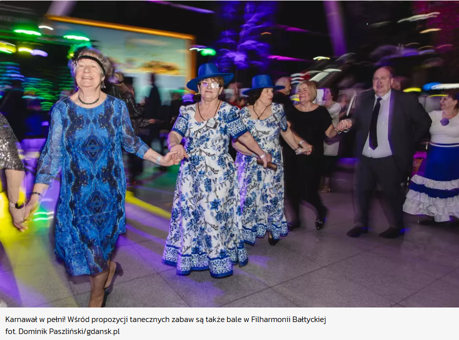 Grupa uśmiechniętych seniorów tańczy w radosnym korowodzie podczas balu karnawałowego w Filharmonii Bałtyckiej. Niektóre uczestniczki mają na sobie pasujące do siebie kwieciste suknie i niebieskie kapelusze.