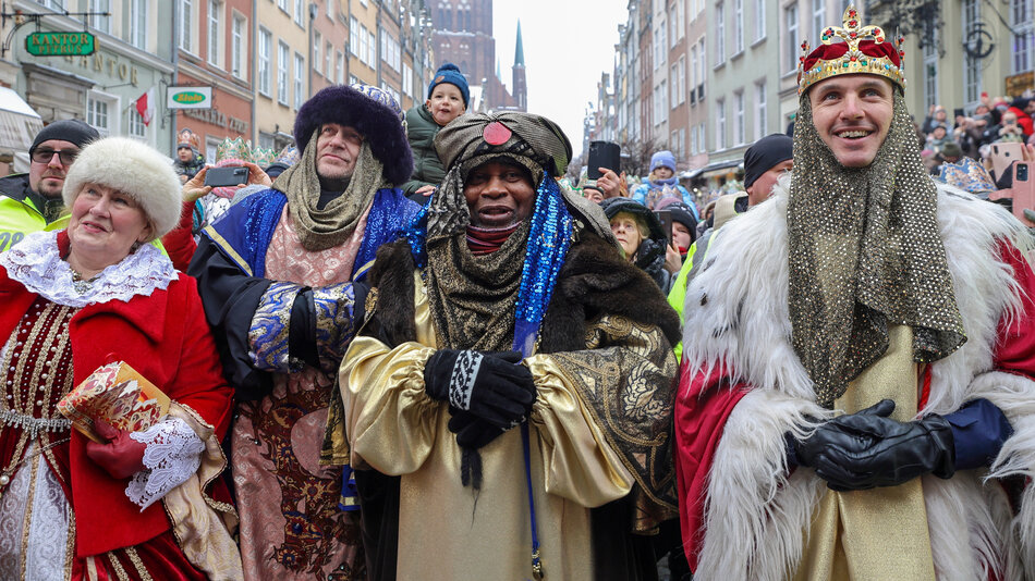  Grupa ludzi ubranych w średniowieczne stroje idąca ulicą. W grupie są cztery osoby, dwóch mężczyzn i dwie kobiety, wszystkie ubrane w kolorowe stroje. Mężczyzna po lewej stronie nosi czerwony płaszcz ze złotymi akcentami i biały kapelusz z czarnym piórem. Trzyma w ręku książkę i wydaje się, że ją czyta. W środku jest mężczyzna ubrany jak król lub szlachcic, ubrany w złotą szatę ze skomplikowanymi wzorami oraz biżuterią. Obok niego inny mężczyźni są ubrani jak żona lub król, również nosi złotą zbroję z klejnotami a na głowie. Po prawej stronie młody mężciec w złotej koronie uśmiecha się i pozuje do zdjęcia. Za nimi można zobaczyć ludzkość idącą po ruchliwej ulicy z budynkami i sklepami po obu stronach. W tle widać ludz