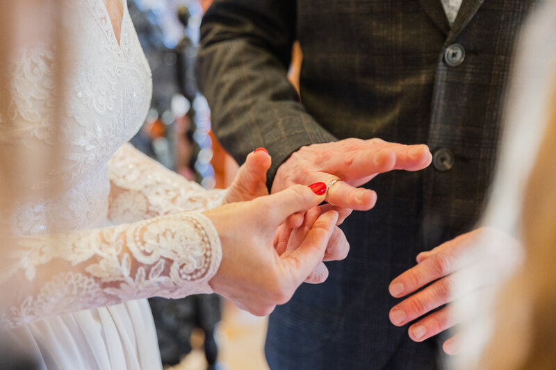 Zbliżenie przedstawia moment nakładania obrączki na palec podczas ceremonii ślubnej. Widoczne dłonie oraz fragmenty stroju podkreślają intymny i uroczysty charakter chwili. 