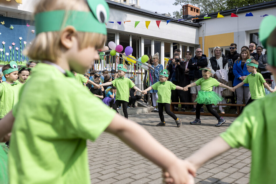 Grupa dzieci ubranych na zielono trzyma się za ręce i tańczy na dziedzińcu podczas plenerowego występu. W tle zgromadzeni dorośli obserwują wydarzenie, a kolorowe dekoracje podkreślają radosny, świąteczny charakter spotkania. 