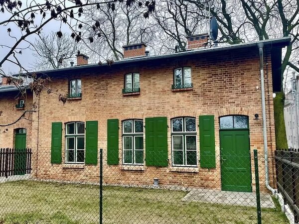 Dwupiętrowy budynek ceglany z pomalowanymi na zielono drzwiami wejściowymi i okiennicami. Przed budynkiem niewielki trawnik otoczony płotem z siatki od frontu i płotem drewnianym po bokach.