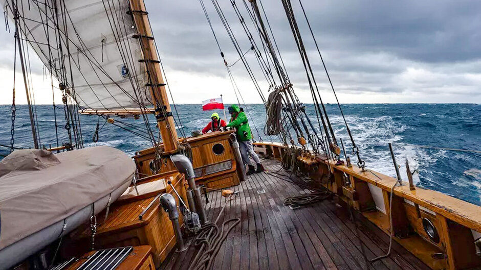 Na zdjęciu widoczny jest pokład żaglowca podczas żeglugi po wzburzonym morzu, z załogą w sztormowych ubraniach pracującą przy takielunku. Ciężkie chmury, przechył jednostki i spienione fale podkreślają trudne warunki pogodowe panujące w trakcie rejsu. 