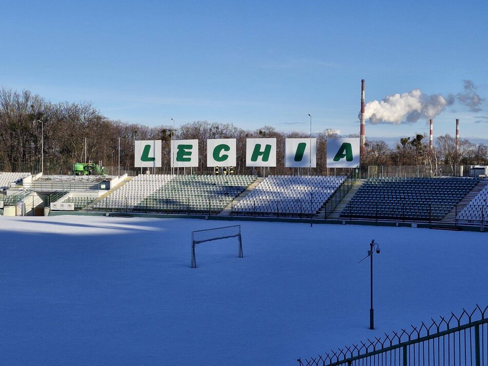 Stadion piłkarski w śniegu, na trybunach widać konstrukcję z zielonym napisem Lechia na białych tablicach.