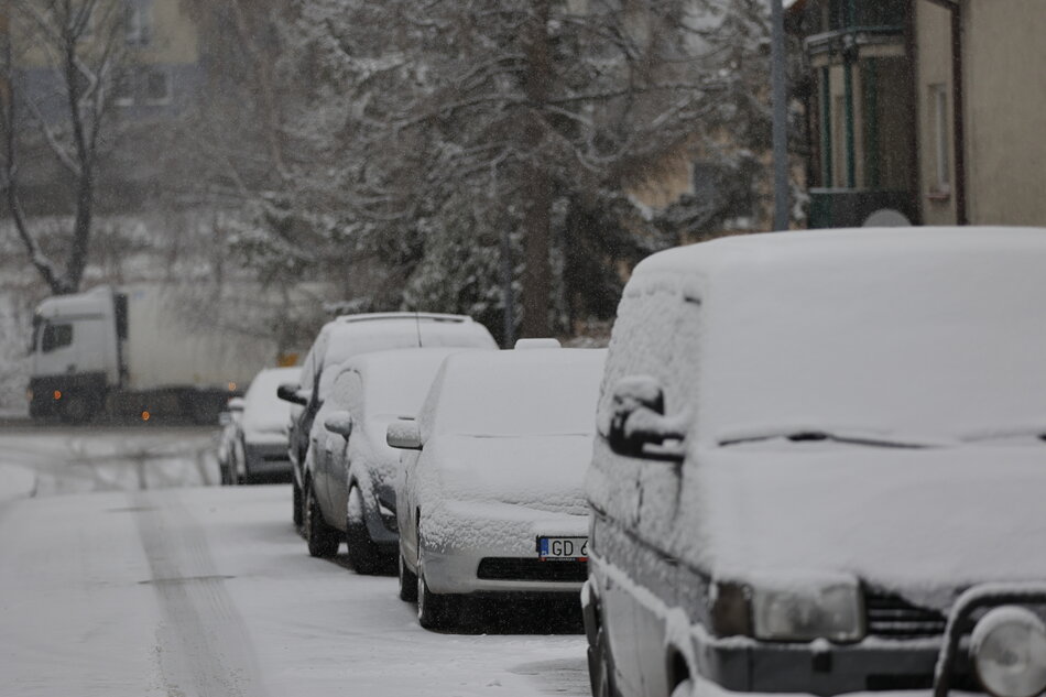 Śnieżna ulica z kilkoma samochodami zaparkowanymi z boku. Auta są pokryte śniegiem