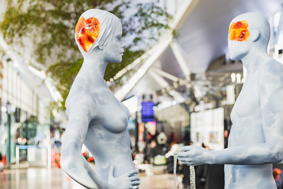 Dwie stylizowane, jasnoniebieskie postacie o ludzkich kształtach stoją naprzeciw siebie w nowoczesnej przestrzeni, przypominającej pasaż lub halę wystawową. W ich głowach widoczne są półprzezroczyste, bursztynowe elementy, które kontrastują z chłodną kolorystyką sylwetek i otoczenia. 