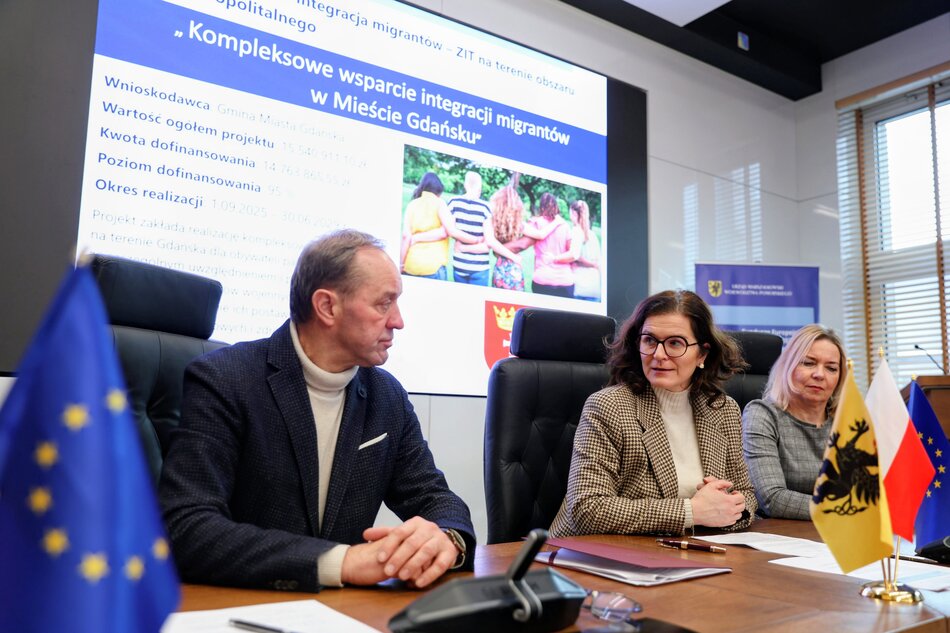 Kobieta i mężczyzna siedzą przy stole o podpisują umowę, za nimi ekran z napisem „Kompleksowe wsparcie integracji migrantów w Mieście Gdańsku”.