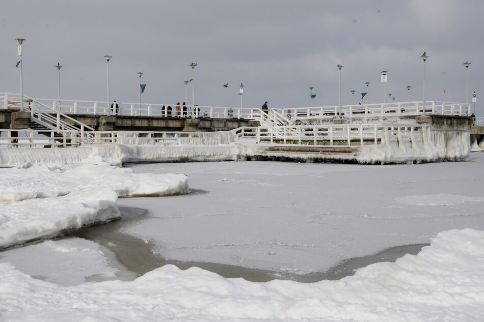  molo na zamarzniętym jeziorze; molo wykonane jest z betonu i ma wiele poziomów z szynami i lekkimi biegunami; na molo chodzi kilka osób, a niebo jest przeklęte; na pierwszym planie jest kilka dużych kawałków lodu i kolców rozrzuconych po lodzie, z których niektóre są częściowo zanurzone w wodzie; po prawej stronie widać, częściowe pokryte lodem.