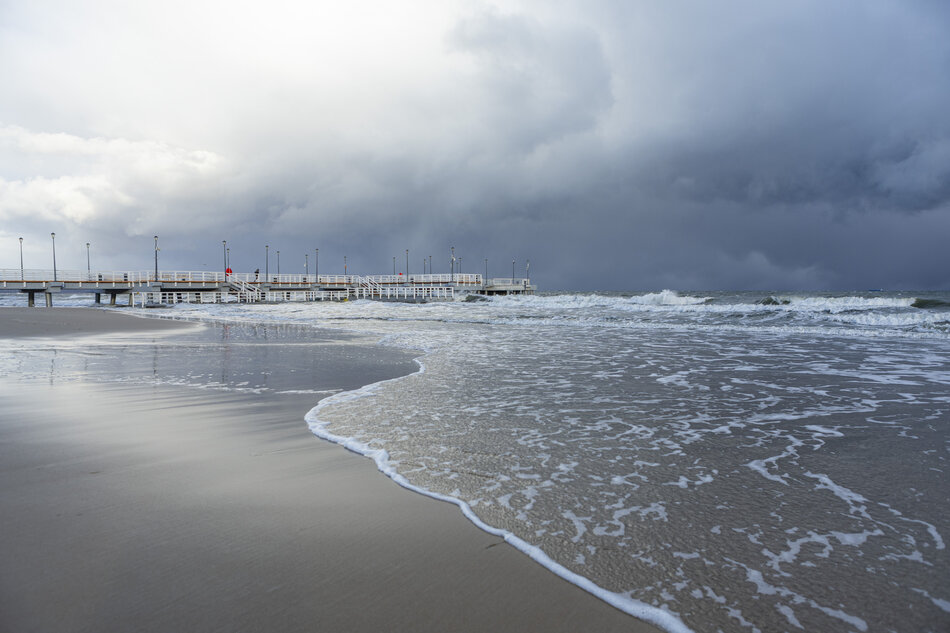  plaża z molo po prawej stronie; niebo jest pochmurne i szare, a woda jest spięta z małymi falami rozbijającymi się na brzeg; po lewej stronie długie drewniane molo z kilkoma biegunami lekkimi rozciągającymi się w odległość; w tle widoczny jest horyzont, z oceanem rozciągającym się przed nim; kilka osób można zobaczyć spacerując po molo, ciesząc się widokiem.