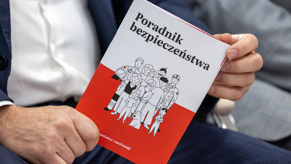 Biało-czerwony dokument o tytule Poradnik Bezpieczeństwa trzymany dwiema rękami przez niewidoczną postać w garniturze