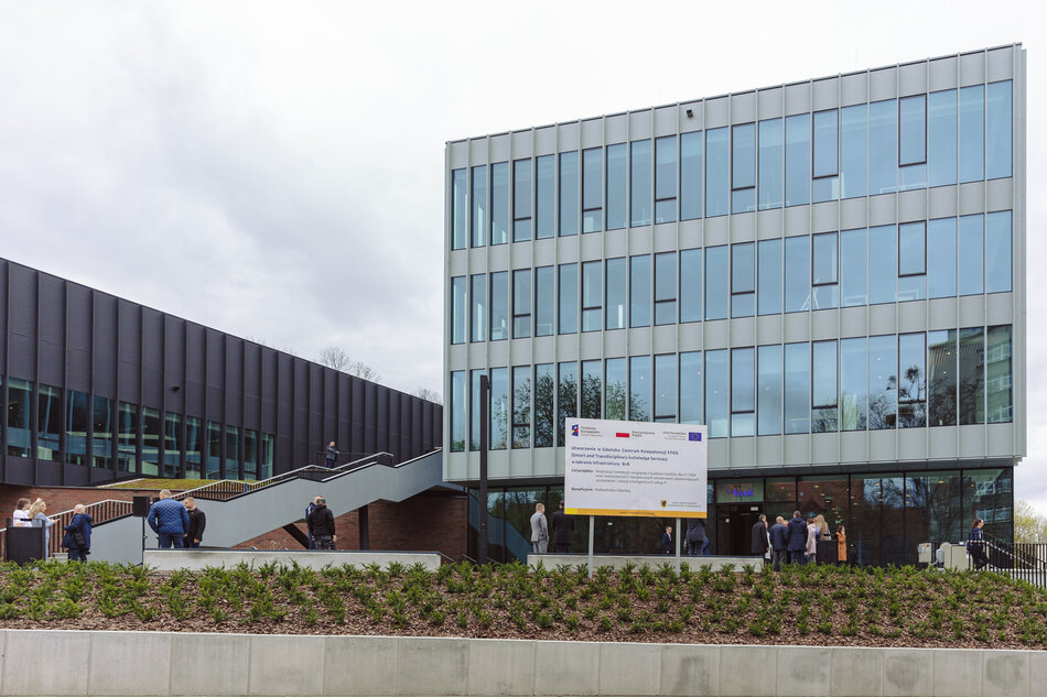 Nowoczesny budynek z dużą szklaną fasadą. Budynek znajduje się na rogu ulicy i jest otoczony betonową ścianą. Na pierwszym planie widać kilka osób spacerujących po chodniku.