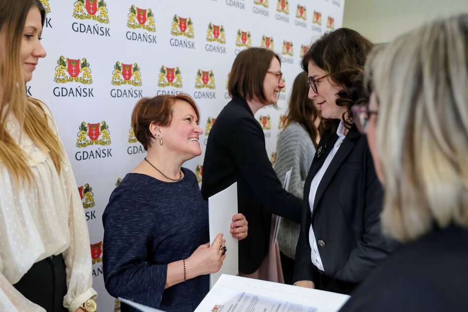 Na zdjęciu widać uczestniczki oficjalnego spotkania prowadzące rozmowę i wymieniające dokumenty na tle ścianki z herbem i logotypem miasta Gdańska. Scena ma formalny charakter i przedstawia moment bezpośrednich gratulacji lub rozmów po zakończeniu części uroczystej. 