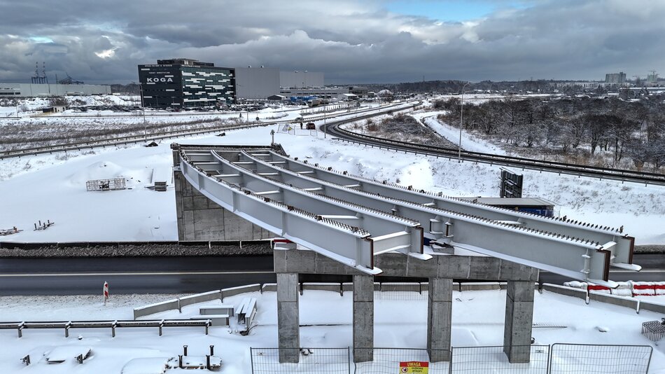 Na zdjęciu przedstawiono konstrukcję wiaduktu drogowego w trakcie realizacji, z widocznymi stalowymi dźwigarami osadzonymi na betonowych podporach. Zimowy krajobraz oraz otaczająca infrastruktura drogowa podkreślają skalę i techniczny charakter inwestycji. 