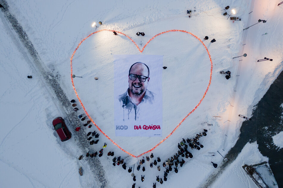 Ujęcie z lotu ptaka przedstawia zgromadzonych ludzi stojących wokół ogromnego serca ułożonego ze świateł na śniegu, w którego centrum znajduje się baner z portretem mężczyzny. Całość ma charakter symbolicznego upamiętnienia i wyrazu solidarności, widocznego zarówno w układzie przestrzennym, jak i obecności uczestników wydarzenia. 
