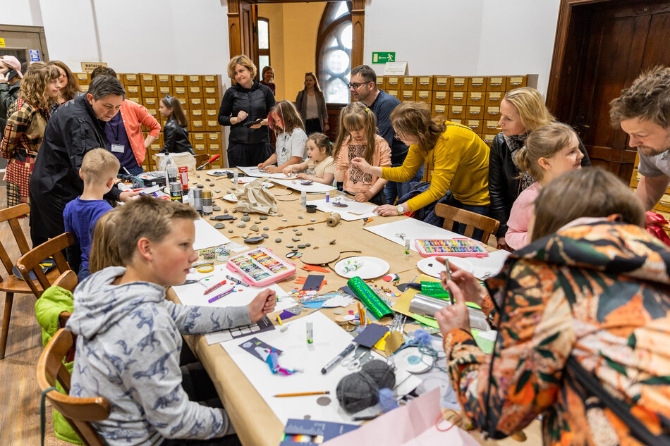 na zdjęciu przy dużym stole siedzą dzieci i kilku dorosłych, na stole widać farbki, kredki, białe kartki do rysowania