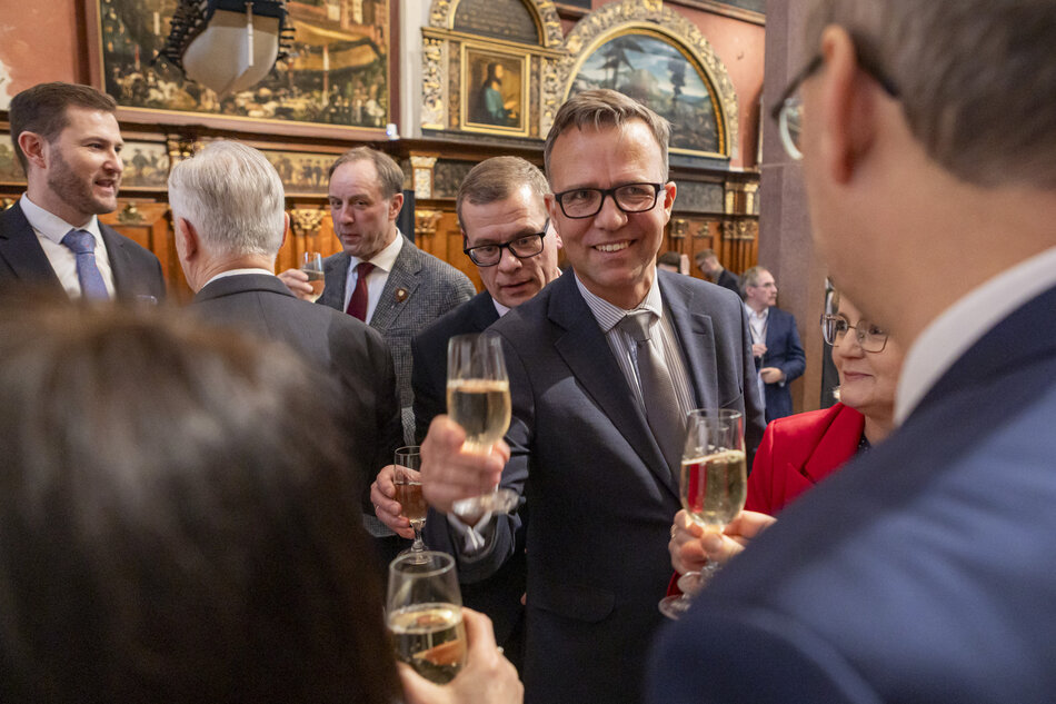 Uczestnicy oficjalnego spotkania w eleganckim wnętrzu wznoszą toast kieliszkami szampana, prowadząc rozmowy w kameralnych grupach. Wydarzenie ma formalny charakter, co podkreślają stroje gości oraz reprezentacyjna oprawa sali z dekoracyjnymi obrazami. 