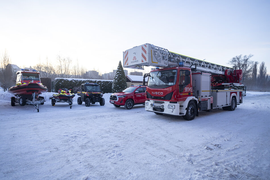 Grupa pojazdów ratowniczych zaparkowanych na pokrytym śniegiem polu. Na zdjęciu znajdują się cztery pojazdy, w tym czerwona ciężarówka strażacka z drabiną z tyłu.