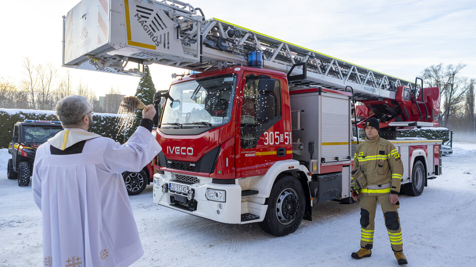  Czerwona ciężarówka Iveco z drabiną przymocowaną do tyłu, ciężarówkę zaparkowano na śnieżnej drodze z drzewami w tle, po lewej stronie znajduje się mężczyzna w białej szacie ze złotym haftem, który wydaje się być kapłanem lub osobą religijną, trzyma mikrofon i rozmawia z strażakiem stojącym przed nim, zarówno mężczyzną, jak i strażakami noszą żółte i czarne mundury z paskami odblaskowymi.