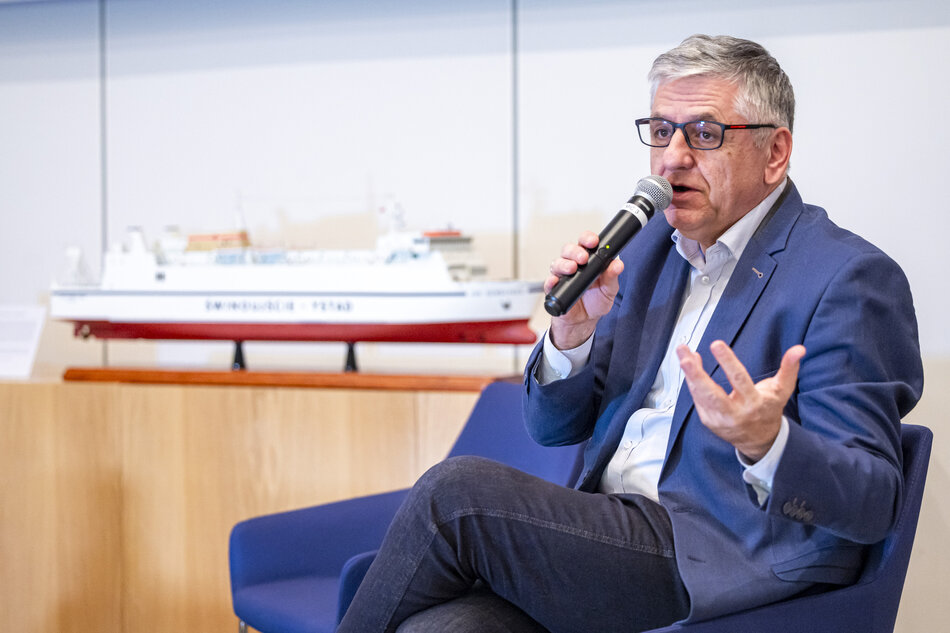 Mężczyzna trzyma mikrofon i prezentuje model statku ustawiony na drewnianej podstawie. Wskazując na detal makiety, prowadzi prelekcję w sali wykładowej. 