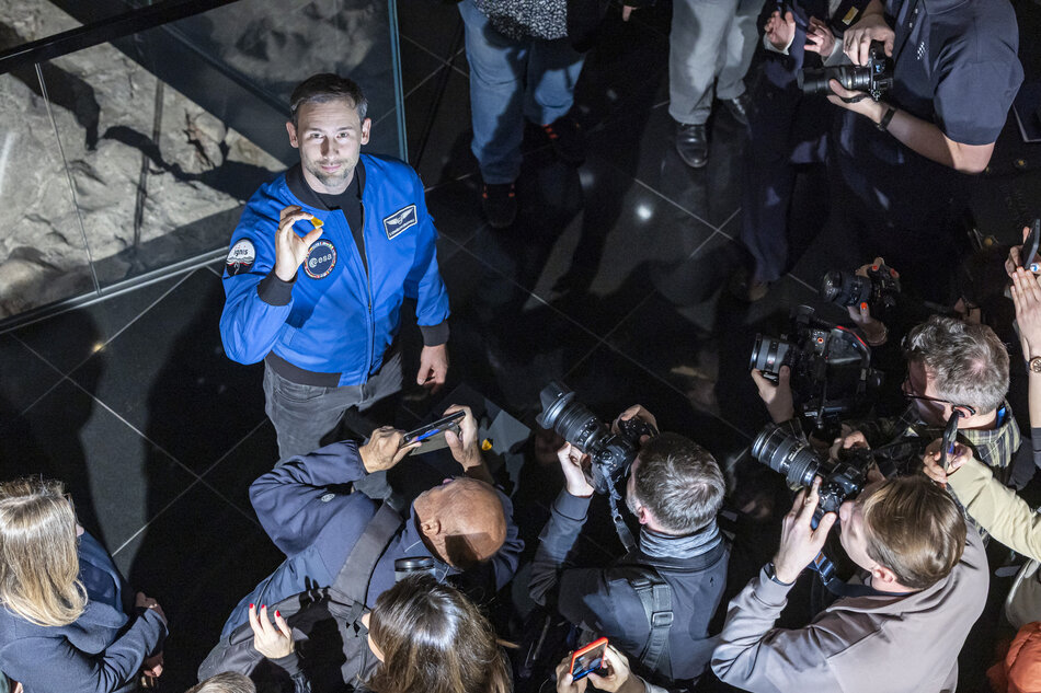 Mężczyzna w stroju astronauty pozuje do zdjęcia pokazując trzymany w palcach kawałek bursztynu. Otaczają go fotoreporterzy. Zdjęcie zrobione z góry, mężczyzna zadziera głowę do góry.