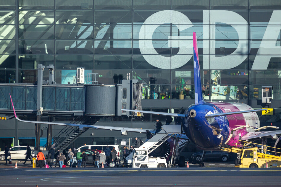 Na zdjęciu widoczny jest samolot zaparkowany przy rękawie pasażerskim na płycie lotniska, z otwartymi drzwiami i podstawionymi schodami. Przed terminalem widać grupę pasażerów oczekujących na wejście na pokład oraz obsługę naziemną przygotowującą maszynę do lotu. 