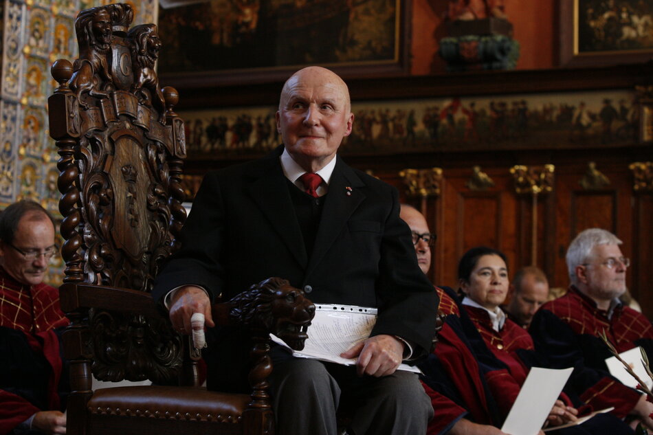Starszy mężczyzna w formalnym, ciemnym stroju siedzi na bogato rzeźbionym, drewnianym fotelu w reprezentacyjnej sali o historycznym wystroju. Wokół niego znajdują się inni uczestnicy ceremonii w jednolitych strojach, a cała scena ma uroczysty, oficjalny charakter. 