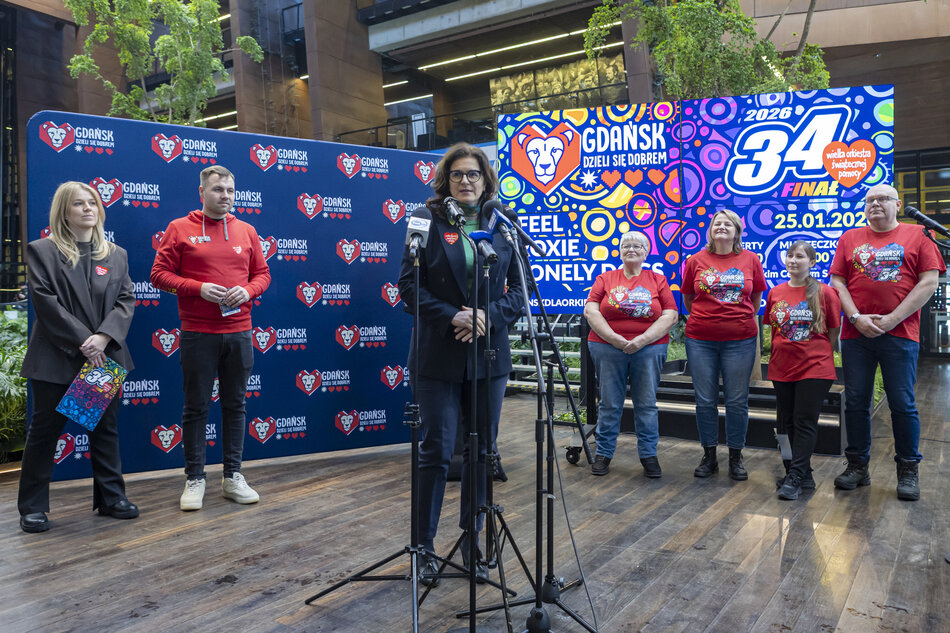 Kobieta mówi do mikrofony, za nią stoi grupa ludzi, wszystko na tle banerów Wielkiej Orkiestry Świątecznej Pomocy.