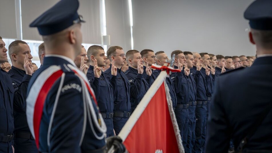 na pierwszym planie po lewej policjant w mundurze trzymający sztandar, w tle stoją w szeregu młodzi policjanci z uniesionymi rękoma na znak ślubowania
