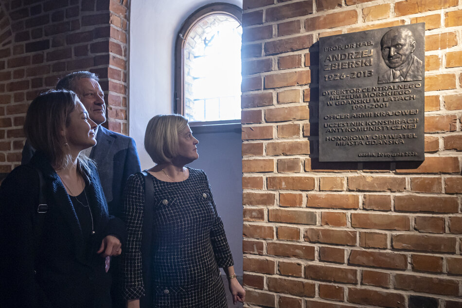 Na zdjęciu widać trzy osoby stojące w ceglanej przestrzeni i przyglądające się pamiątkowej tablicy zamontowanej na ścianie. Tablica zawiera inskrypcję poświęconą Andrzejowi Zbierskiemu, a całość doświetla światło wpadające przez niewielkie okno. 