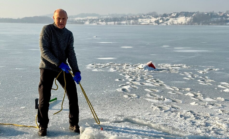 Mężczyzna stoi na zamarzniętym jeziorze i przy pomocy liny wyciąga z przerębla fragment lodu. W tle widać ośnieżony brzeg oraz ślady prowadzące do miejsca pracy na lodzie. 