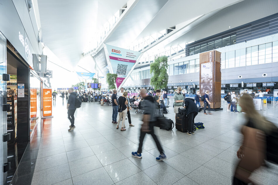 Na zdjęciu widać przestronną halę terminala lotniczego z licznymi podróżnymi przemieszczającymi się między stanowiskami odprawy i punktami usługowymi. Jasne wnętrze z wysokim sufitem, dużymi przeszkleniami i tablicami informacyjnymi podkreśla nowoczesny charakter lotniska. 