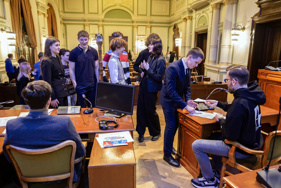 Młodzi ludzie stoją i siedzą, rozmawiają i śmieją się w dużej reprezentacyjnej sali w starym stylu.