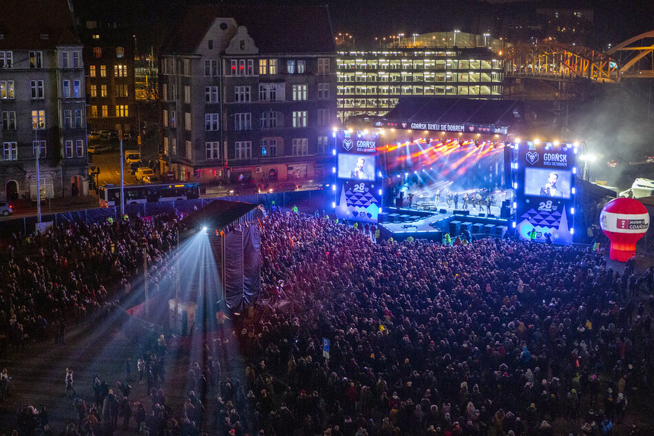 duży tłum ludzi zebranych na miejskim placu w nocy. Plac otoczony jest budynkami i w tle znajduje się most. W centrum placu znajdują się oświetlona duża scena