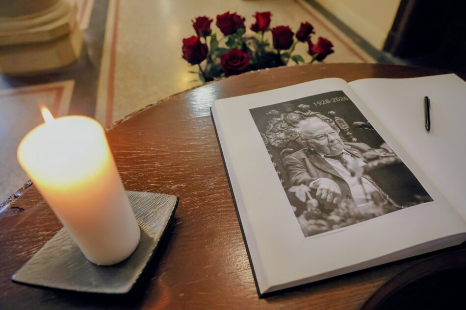 Na drewnianym pulpicie spoczywa księga pamiątkowa otwarta na stronie z czarno-białym portretem mężczyzny oraz datami 1928–2026, obok której leży długopis. Obok księgi pali się biała świeca, a w tle widoczny jest bukiet czerwonych róż, tworzący nastrojową, żałobną kompozycję. 