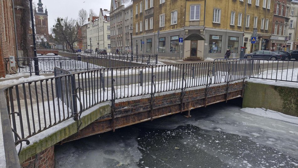 Zaśnieżona, brukowana jezdnia prowadzi przez niewielki most otoczony metalowymi barierkami, pomiędzy kamienicami o zróżnicowanej architekturze. Zimowa sceneria z resztkami śniegu na chodnikach i dachach budynków podkreśla spokojny, miejski charakter miejsca. 