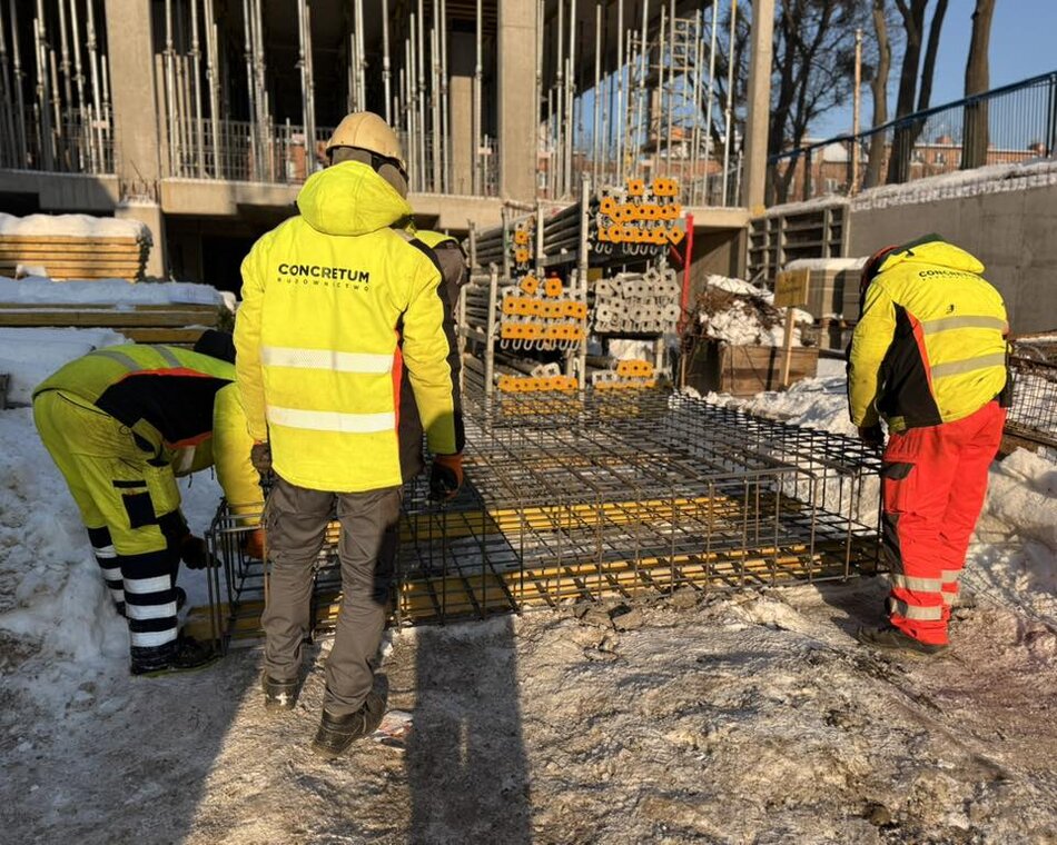 na zdjęciu robotnicy budowlani w jaskrawych kombinezonach, przed nimi budowany budynek
