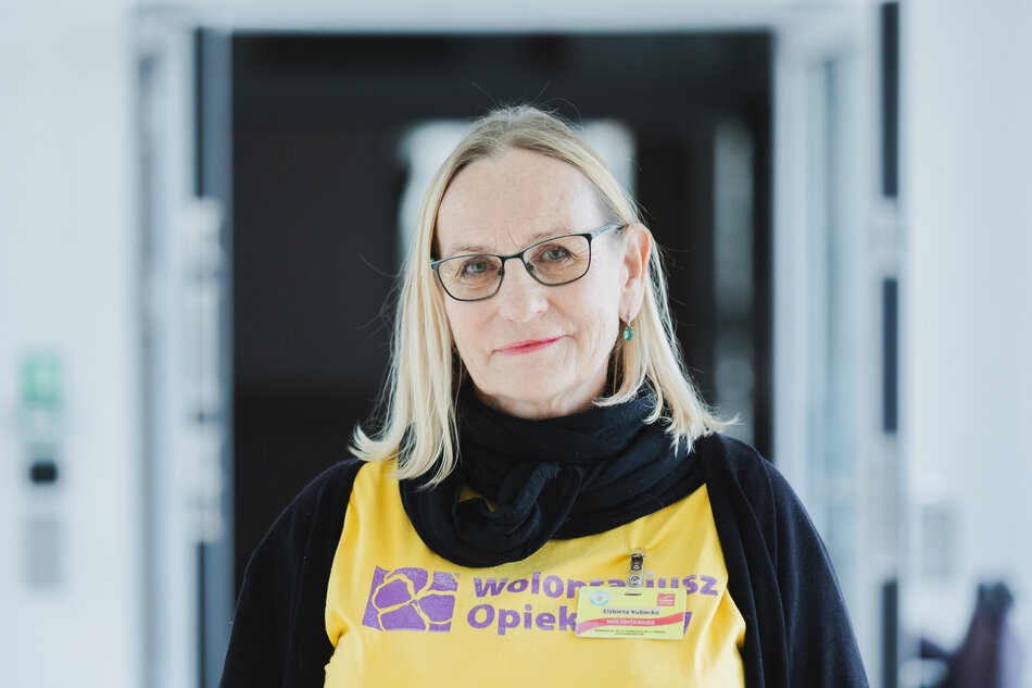 Wolontariuszka w żółtej koszulce stoi przy łóżku osoby leżącej i trzyma jej dłoń, okazując wsparcie i bliskość. Scena rozgrywa się w spokojnym wnętrzu pokoju, którego wystrój i światło podkreślają kameralny, opiekuńczy charakter sytuacji. 