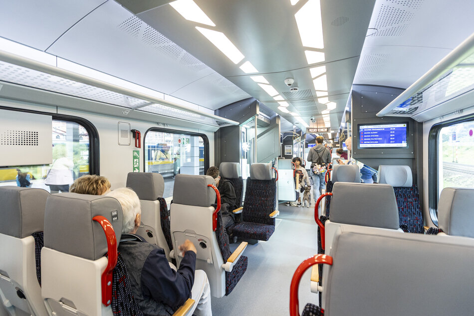 Wnętrze nowoczesnego pociągu z jasnym oświetleniem, rzędami siedzeń i pasażerami zajmującymi miejsca lub stojącymi w przejściu. Widoczne ekrany informacyjne, przestrzeń dla podróżnych oraz przeszklone drzwi podkreślają funkcjonalny i współczesny charakter wagonu. 