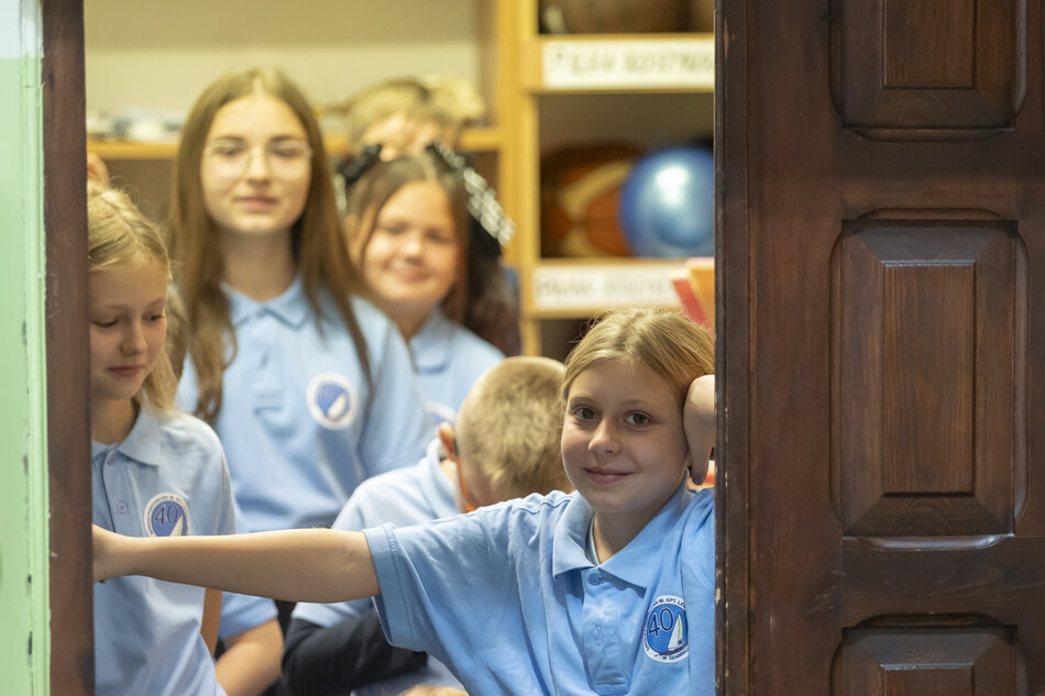 Grupa dzieci w jasnoniebieskich koszulkach stoi w drzwiach sali, a jedno z nich opiera się o futrynę i spogląda w stronę obiektywu. W tle widoczne są półki z pomocami, co sugeruje szkolne wnętrze i codzienną atmosferę zajęć. 